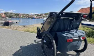 En dubbel joggingvagn står vid en svensk kust med röda hus och båtar i bakgrunden.