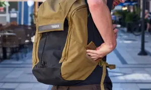 En person med en Thule ryggsäck i kamelfärg och svart. Ryggsäcken har ett Thule-märke och en person stoppar in handen i en ficka.