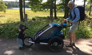 En kvinna med ryggsäck och ett barn vid en åkpåse Thule Chariot på en grusväg i skogen.