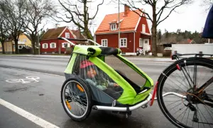 Ett barn sitter i en grön och grå spectra cykelvagn på en regnvåt cykelbana. Röda hus syns i bakgrunden.