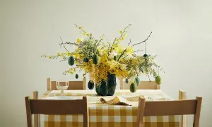 En vacker påskris inspiration med gula blommor och gröna ägg som hänger från grenarna. Duken är gulrutig.
