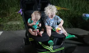 Two young children sit in a double stroller, eating corn from cups.