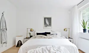 Sovrum med stor säng, vita sängkläder, mörka kuddar, guldig lampa och konst. Enkel inredning som skapar lugn, perfekt för feng shui sovrum.