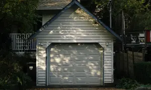 Ett vitt garage med en takås och en garageport. Trädskuggor faller över porten. Tänker du på bygga garage kostnad?