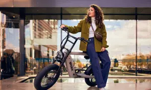 Kvinna med lockigt hår sitter på en grå elcykel med tjocka däck. Hon bär en grön jacka och jeans. Cykeln är ett perfekt exempel på fatbike tillbehör för stadspendlare.