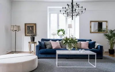 Blå soffa inspiration: En elegant salong med en djupblå sammetssoffa, prydd med kuddar i rosa och grått. Ett modernt soffbord och en stor golvlampa kompletterar rummet.