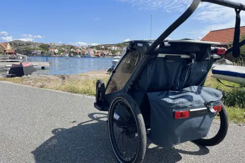 En dubbel joggingvagn står vid en svensk kust med röda hus och båtar i bakgrunden.