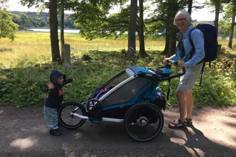 En kvinna med ryggsäck och ett barn vid en åkpåse Thule Chariot på en grusväg i skogen.