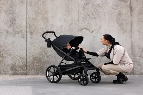Glad baby i svart cityvagn, en kvinna böjer sig ner för att leka med barnet.