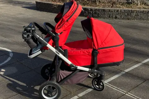 En röd Thule sleek syskonvagn står på en stenlagd uteplats. Vagnen har en liggdel och en sittdel, båda i rött.
