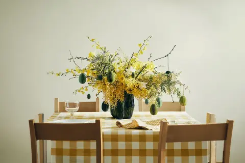 En vacker påskris inspiration med gula blommor och gröna ägg som hänger från grenarna. Duken är gulrutig.