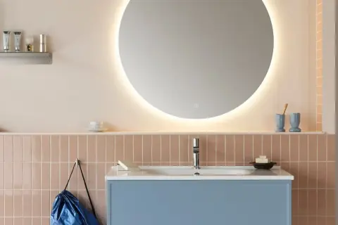 Ett modernt badrum med en blå kommod, rosa kakel och en stor, upplyst rund spegel. En diskret inredning med speglar som skapar rymd.