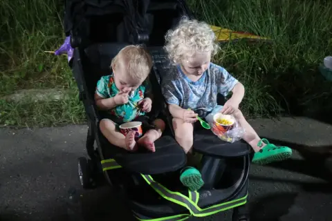 Two young children sit in a double stroller, eating corn from cups.