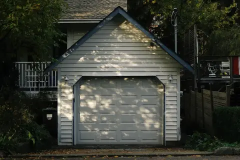 Ett vitt garage med en takås och en garageport. Trädskuggor faller över porten. Tänker du på bygga garage kostnad?