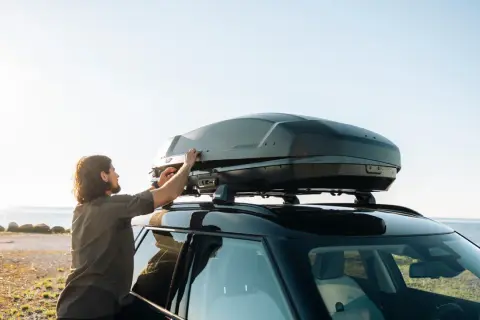Man justerar en Thule lastbox på taket av en bil vid havet.