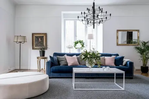 Blå soffa inspiration: En elegant salong med en djupblå sammetssoffa, prydd med kuddar i rosa och grått. Ett modernt soffbord och en stor golvlampa kompletterar rummet.