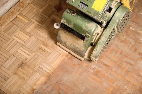 En golvslipmaskin står på ett parkettgolv. Dammet visar var man har börjat slipa golvet.