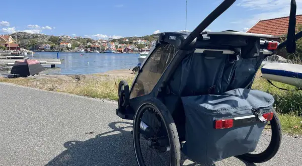 En dubbel joggingvagn står vid en svensk kust med röda hus och båtar i bakgrunden.