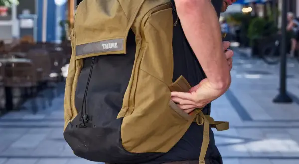 En person med en Thule ryggsäck i kamelfärg och svart. Ryggsäcken har ett Thule-märke och en person stoppar in handen i en ficka.