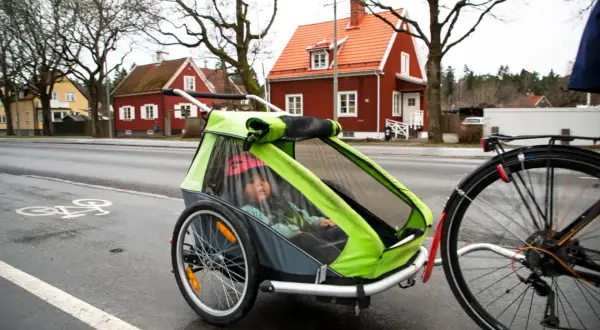 Ett barn sitter i en grön och grå spectra cykelvagn på en regnvåt cykelbana. Röda hus syns i bakgrunden.