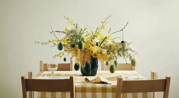 En vacker påskris inspiration med gula blommor och gröna ägg som hänger från grenarna. Duken är gulrutig.