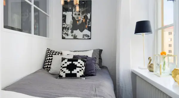 Stilren inredning litet sovrum med Audrey Hepburn-poster, grå sängkläder och grafiska kuddar. En golvlampa och en blomma på fönsterbrädan.