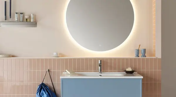 Ett modernt badrum med en blå kommod, rosa kakel och en stor, upplyst rund spegel. En diskret inredning med speglar som skapar rymd.
