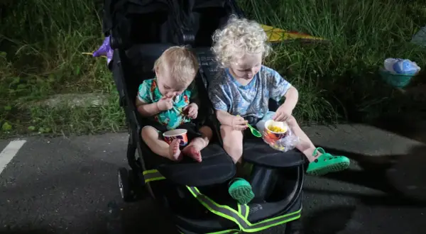 Two young children sit in a double stroller, eating corn from cups.
