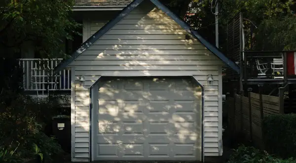 Ett vitt garage med en takås och en garageport. Trädskuggor faller över porten. Tänker du på bygga garage kostnad?