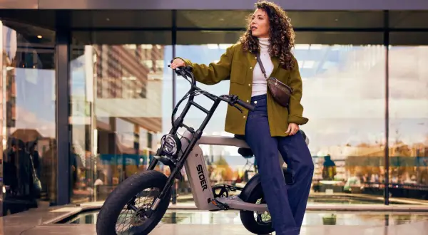Kvinna med lockigt hår sitter på en grå elcykel med tjocka däck. Hon bär en grön jacka och jeans. Cykeln är ett perfekt exempel på fatbike tillbehör för stadspendlare.