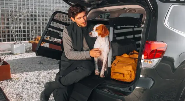 Man and dog sitting in the open trunk of a car with a dog crate and luggage.