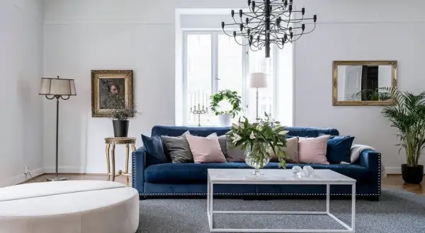 Blå soffa inspiration: En elegant salong med en djupblå sammetssoffa, prydd med kuddar i rosa och grått. Ett modernt soffbord och en stor golvlampa kompletterar rummet.