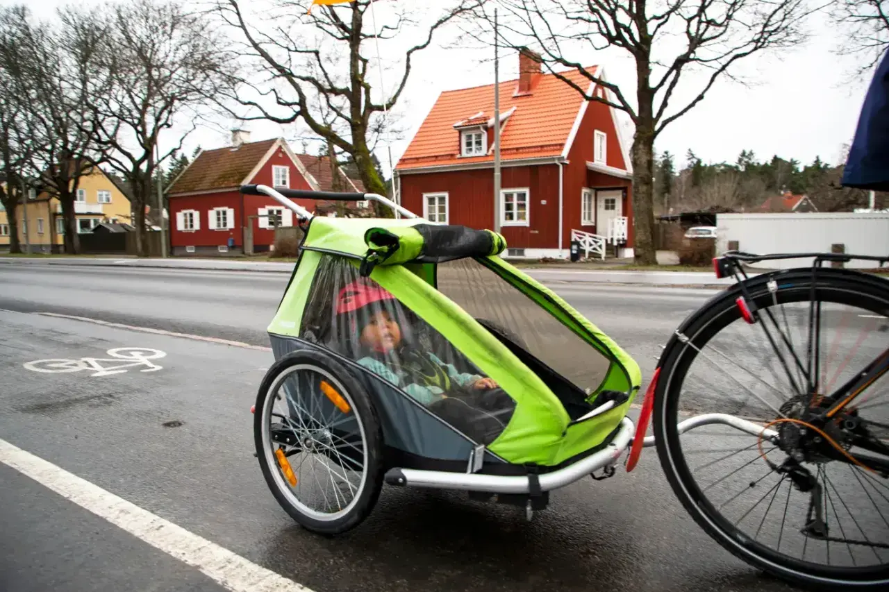 Ett barn sitter i en grön och grå spectra cykelvagn på en regnvåt cykelbana. Röda hus syns i bakgrunden.