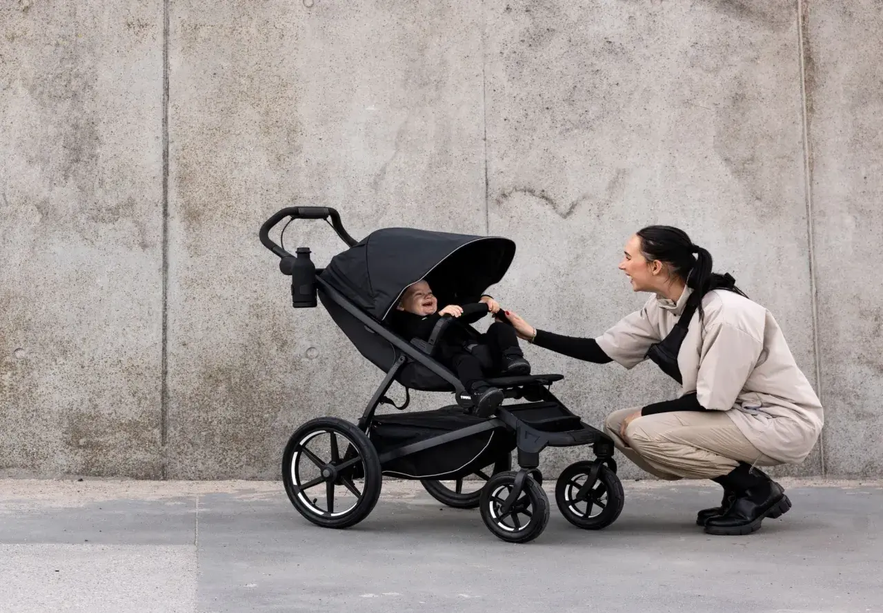 Glad baby i svart cityvagn, en kvinna böjer sig ner för att leka med barnet.