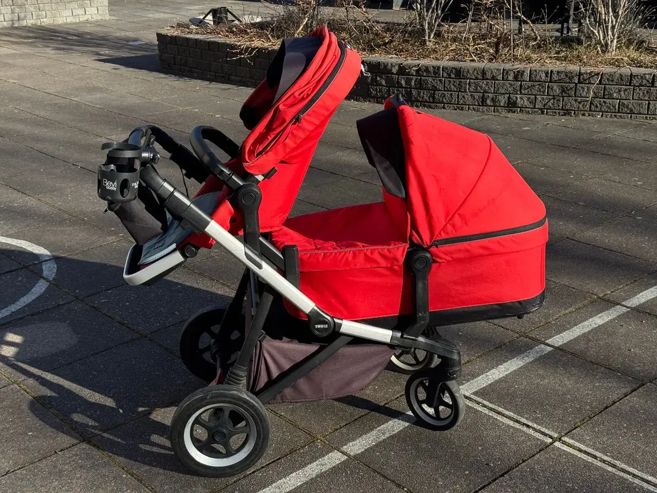 En röd Thule sleek syskonvagn står på en stenlagd uteplats. Vagnen har en liggdel och en sittdel, båda i rött.