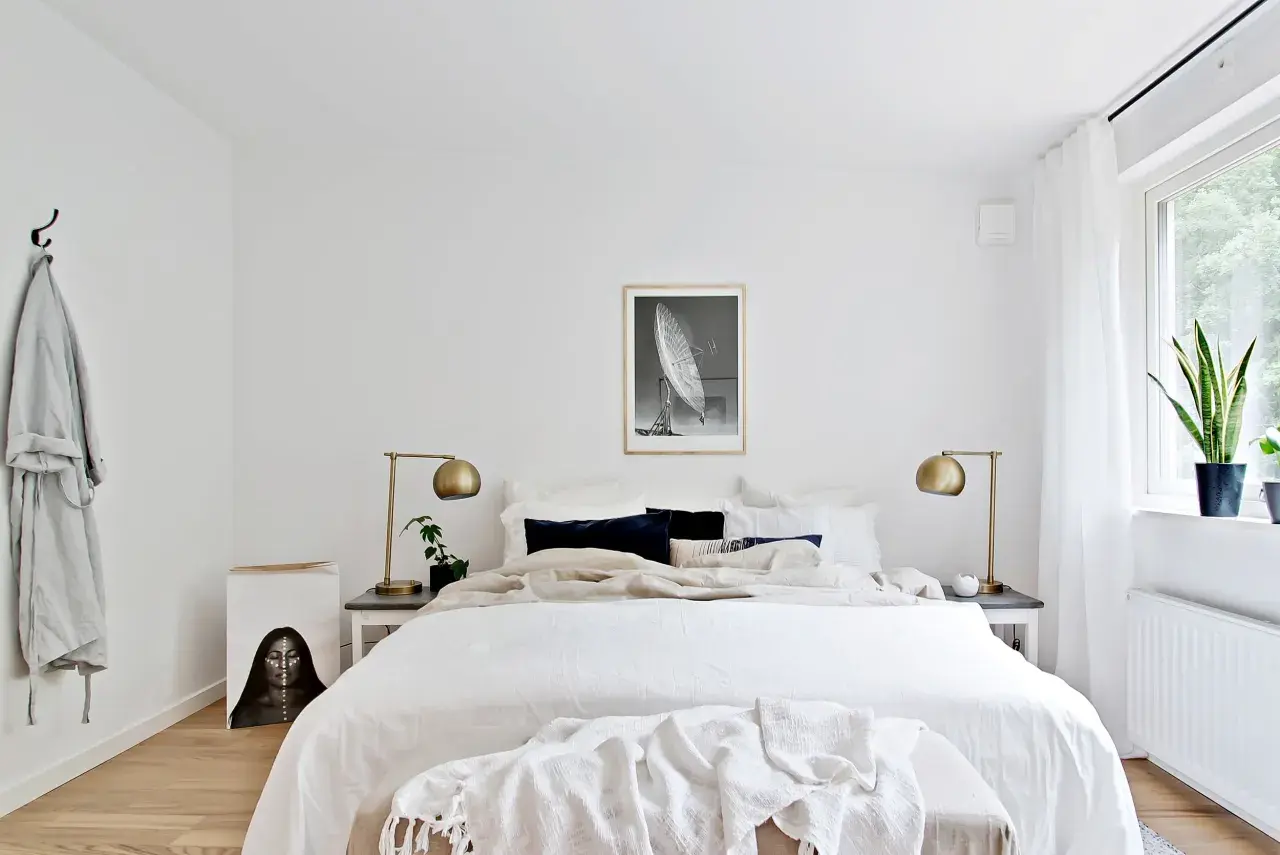 Sovrum med stor säng, vita sängkläder, mörka kuddar, guldig lampa och konst. Enkel inredning som skapar lugn, perfekt för feng shui sovrum.