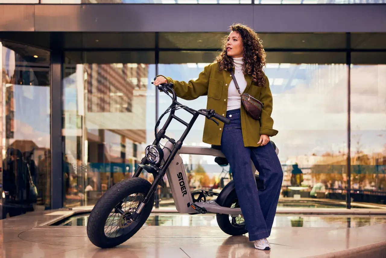 Kvinna med lockigt hår sitter på en grå elcykel med tjocka däck. Hon bär en grön jacka och jeans. Cykeln är ett perfekt exempel på fatbike tillbehör för stadspendlare.