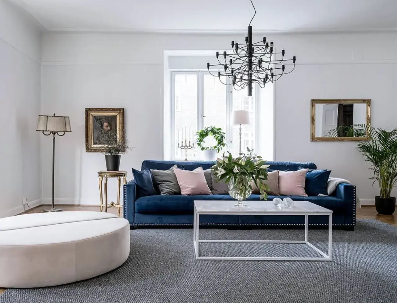 Blå soffa inspiration: En elegant salong med en djupblå sammetssoffa, prydd med kuddar i rosa och grått. Ett modernt soffbord och en stor golvlampa kompletterar rummet.