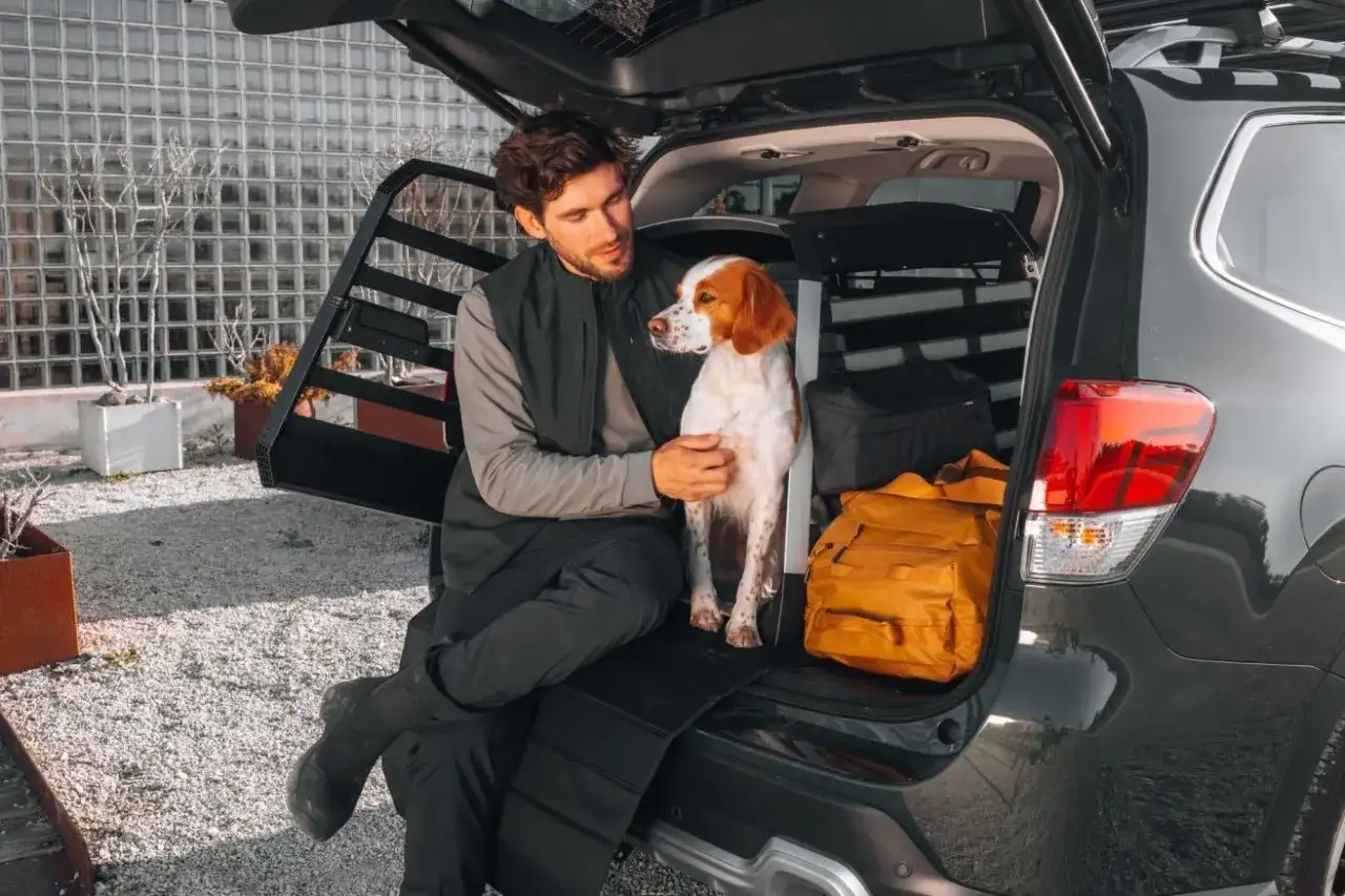 Man and dog sitting in the open trunk of a car with a dog crate and luggage.