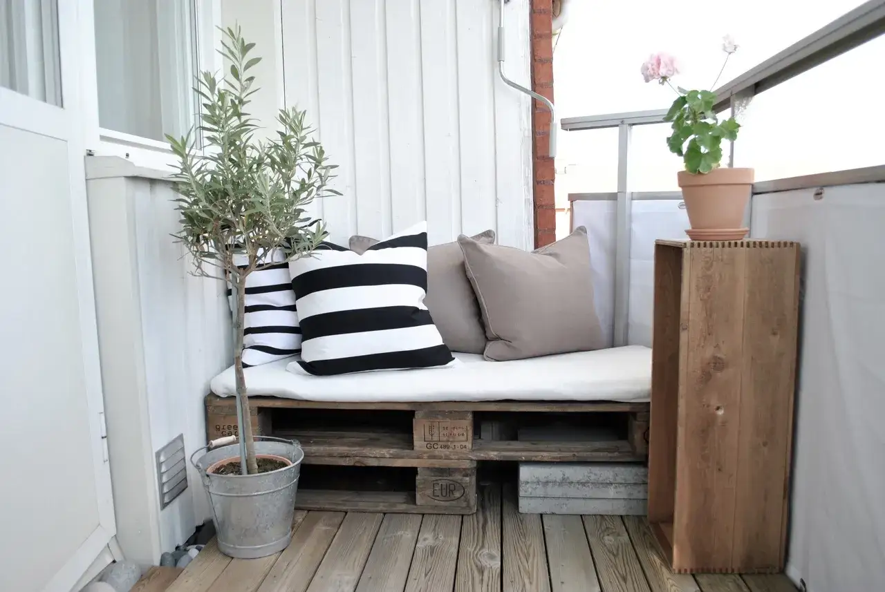 Mysig balkong med en soffa byggd av lastpallar, prydd med randiga och enf&auml;rgade kuddar. En olivtr&auml;d i en zinkhink ger gr&ouml;nska.