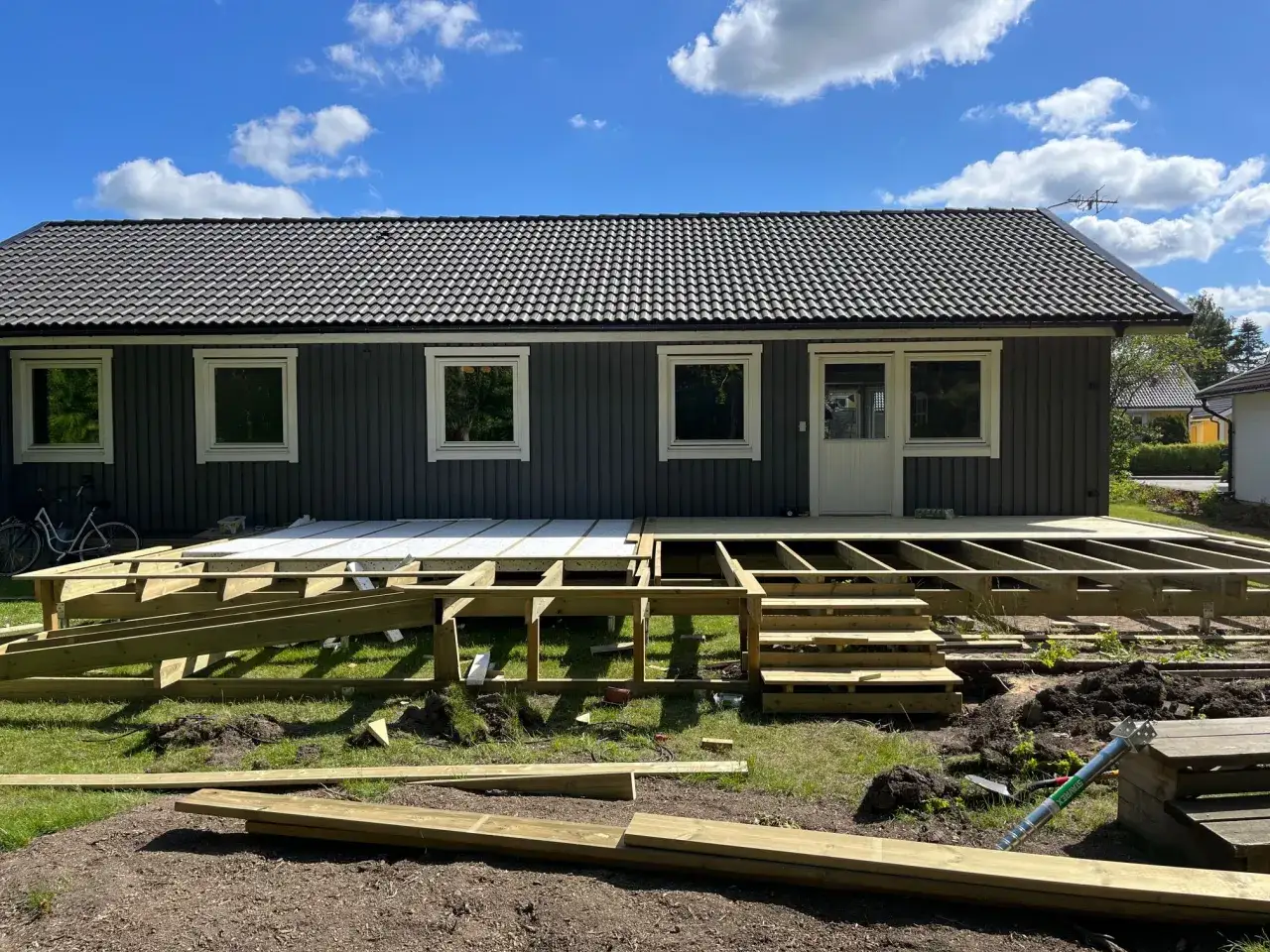 Bygga altan: Tr&auml;konstruktion f&ouml;r en ny altan tar form framf&ouml;r ett m&ouml;rkt tr&auml;hus. Solen skiner p&aring; en bl&aring; himmel med moln.