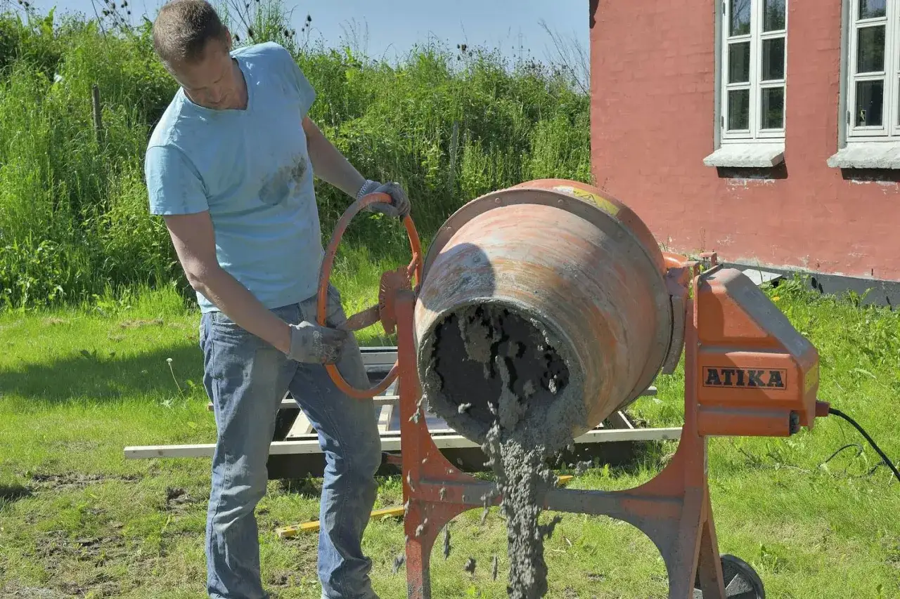 Man h&auml;ller ut f&auml;rdigblandad betong ur en cementblandare. Han &auml;r utomhus vid ett r&ouml;tt hus.