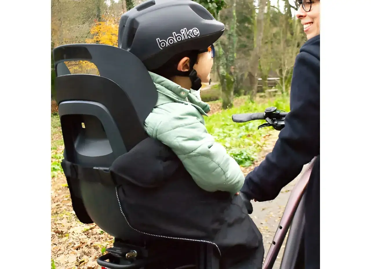 En person med glasögon ler medan ett barn sitter i en cykelbarnstol, redo för en tur i skogen.