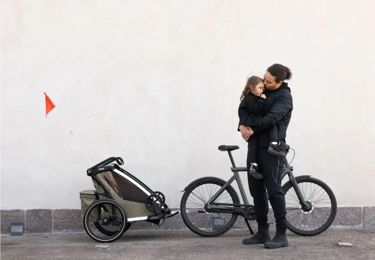 Pappa med barn i famnen bredvid cykel och barnvagn. Thule tillbeh&ouml;r f&ouml;r aktiva familjer.
