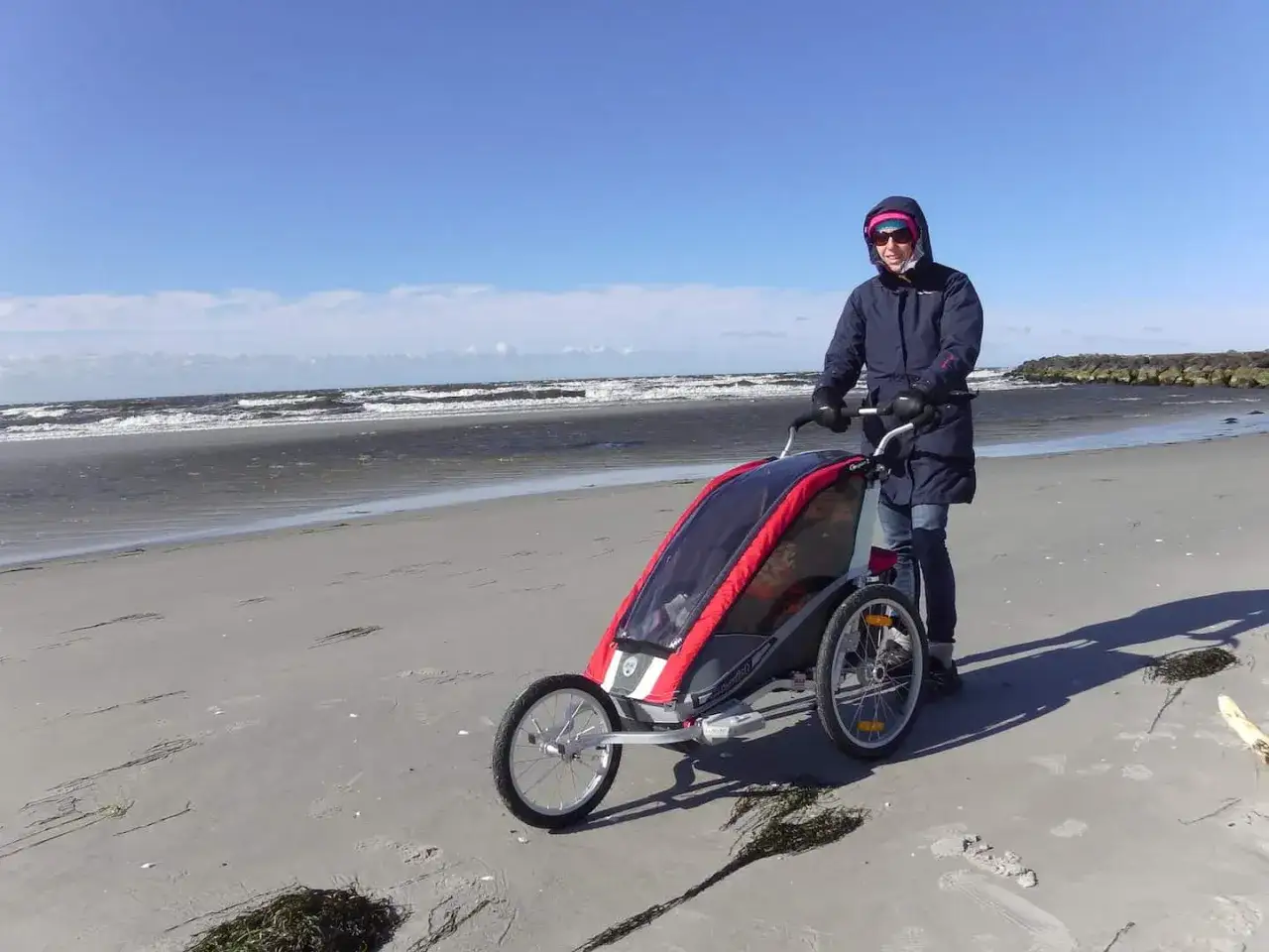 En person knuffar en multisportvagn p&aring; en sandstrand med havet i bakgrunden.