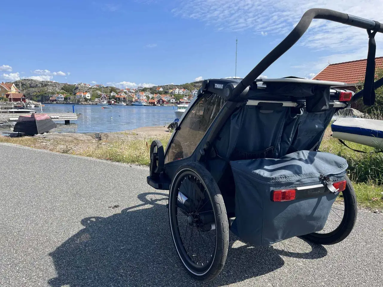 En Thule Chariot barnvagn med &aring;kp&aring;se st&aring;r vid en svensk kust med r&ouml;da hus och b&aring;tar i bakgrunden.