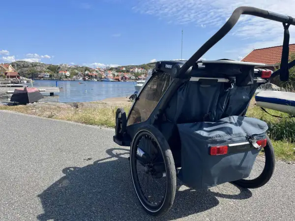 En dubbel joggingvagn står vid en svensk kust med röda hus och båtar i bakgrunden.