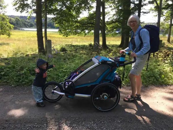 En kvinna med ryggsäck och ett barn vid en åkpåse Thule Chariot på en grusväg i skogen.