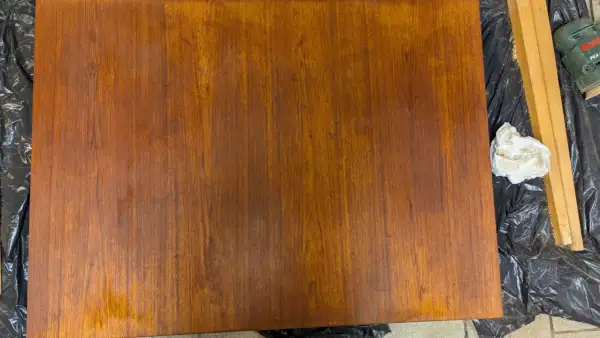 Förbereder för att slipa teakbord. En bit trä ligger på svart plast, bredvid en slipmaskin och en pappersbit.