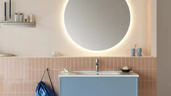 Ett modernt badrum med en blå kommod, rosa kakel och en stor, upplyst rund spegel. En diskret inredning med speglar som skapar rymd.